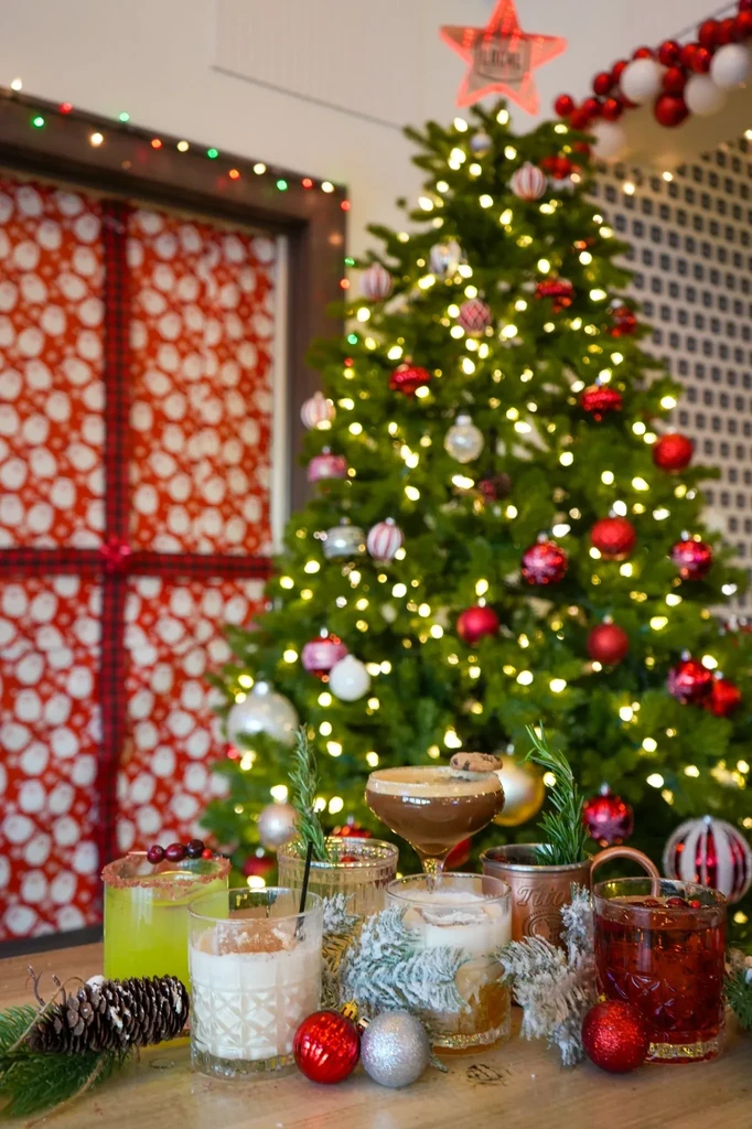 Five festive cocktails on a table with Christmas decorations and a tree.