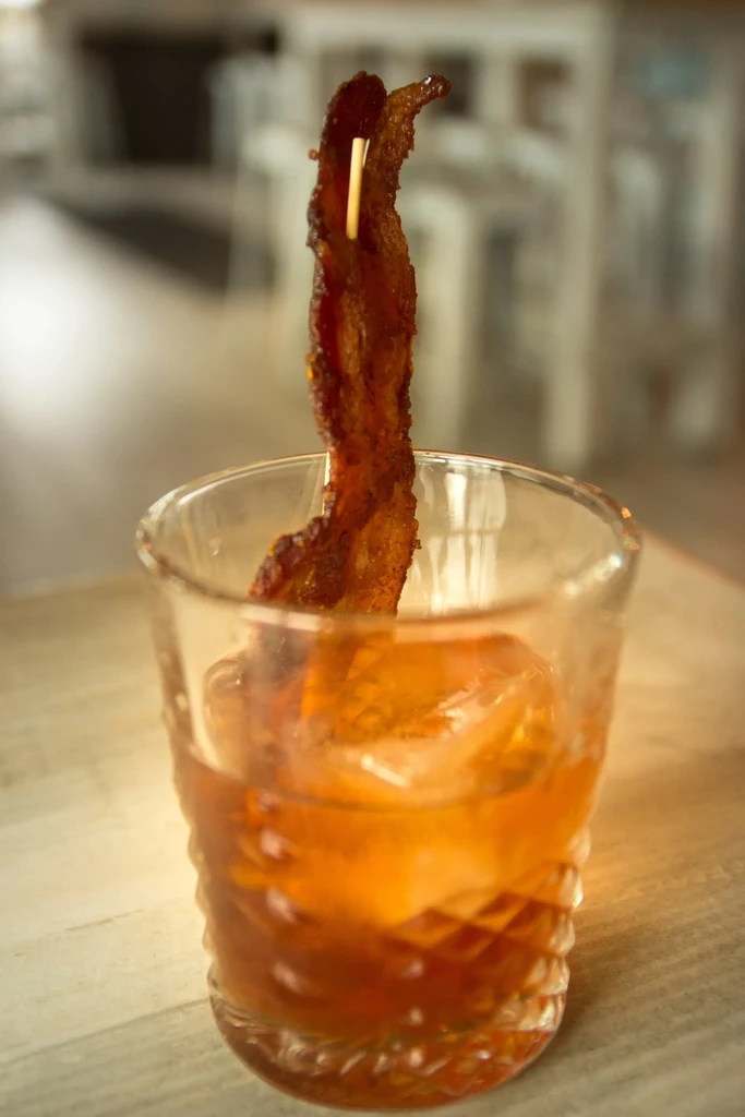 Crispy bacon strip garnishes an amber whiskey cocktail in a textured glass.