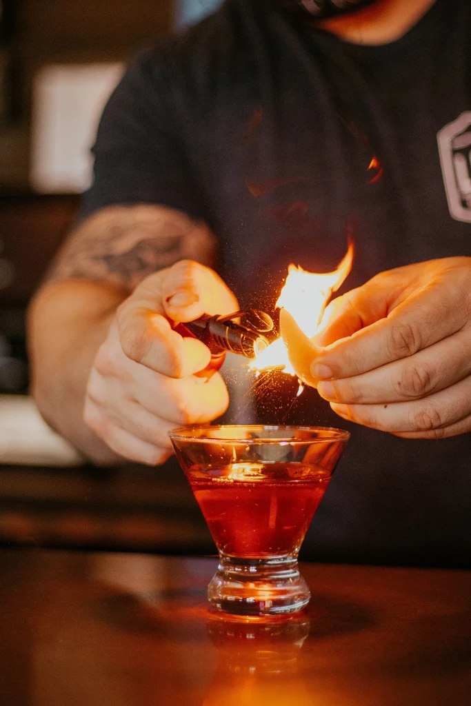 Bartender flares orange peel over cocktail in martini glass, fiery burst.
