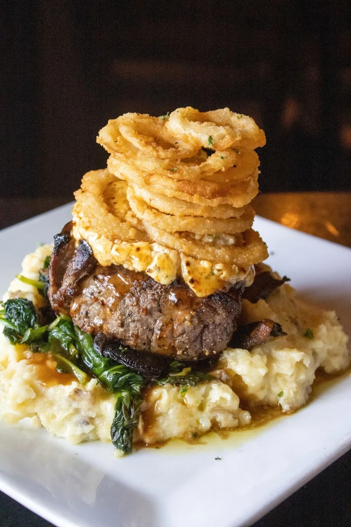 A tall stack of golden onion rings on a steak and mashed potatoes.