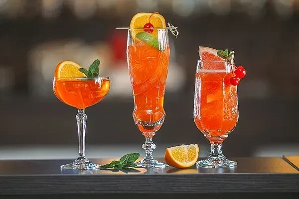 Three orange cocktails garnished with fruit and mint on a bar.