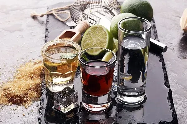 Three shot glasses of different alcoholic drinks with limes, sugar, and ice.