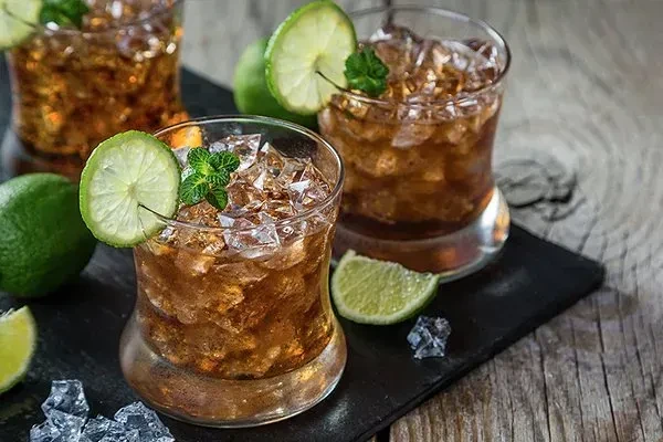 Three dark drinks with ice, lime, and mint on a wooden table.