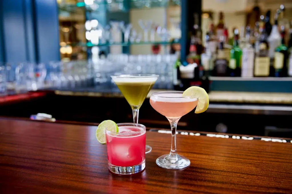 Three cocktails on a dark wood bar with liquor bottles behind.