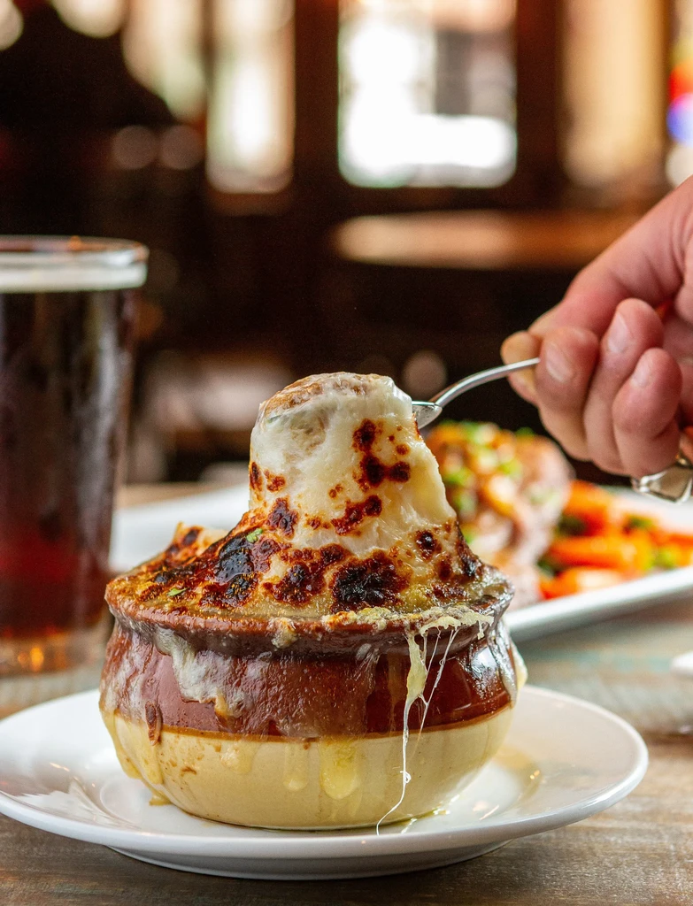 A hand uses a spoon to scoop cheesy French onion soup from a bowl.