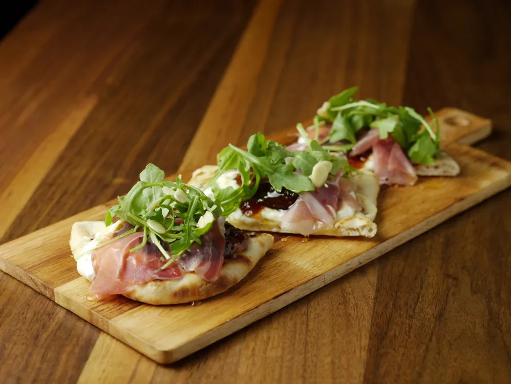 Three flatbread appetizers with prosciutto, arugula, and cheese on a wooden board.