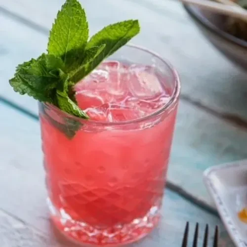 Pink cocktail with ice and mint on a light blue table.