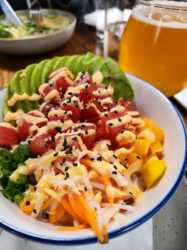 A poke bowl with tuna, avocado, vegetables, sauce, and a beer.