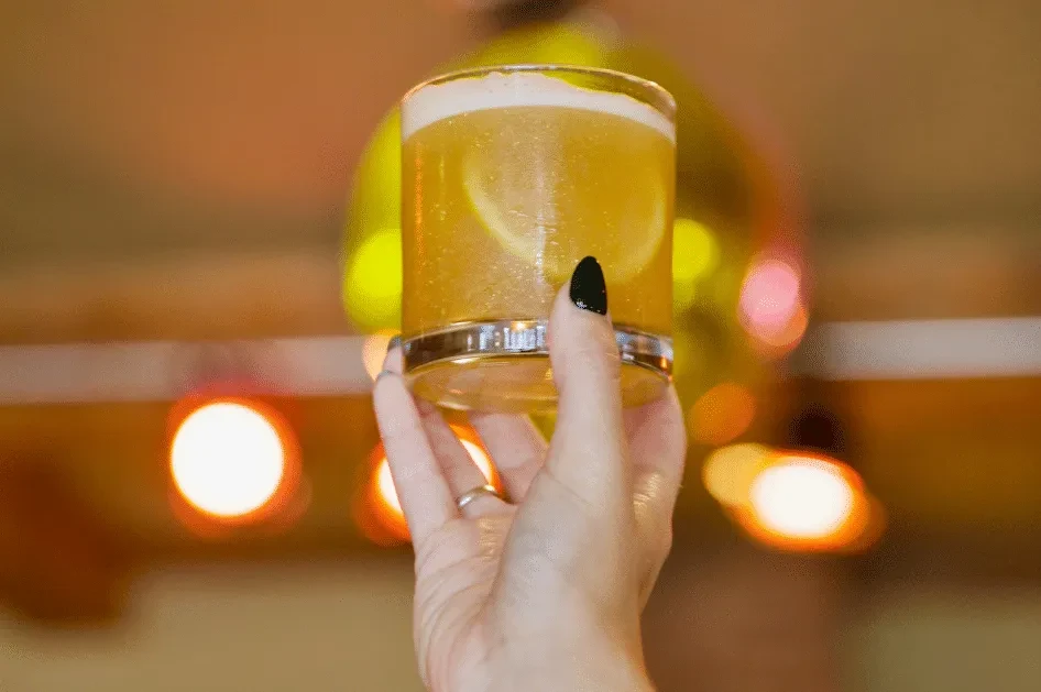 Hand holding a shimmering yellow cocktail with lemon slices and foam.