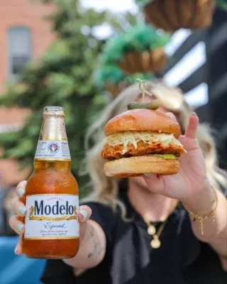 A person holds a Modelo beer and a chicken sandwich with slaw.