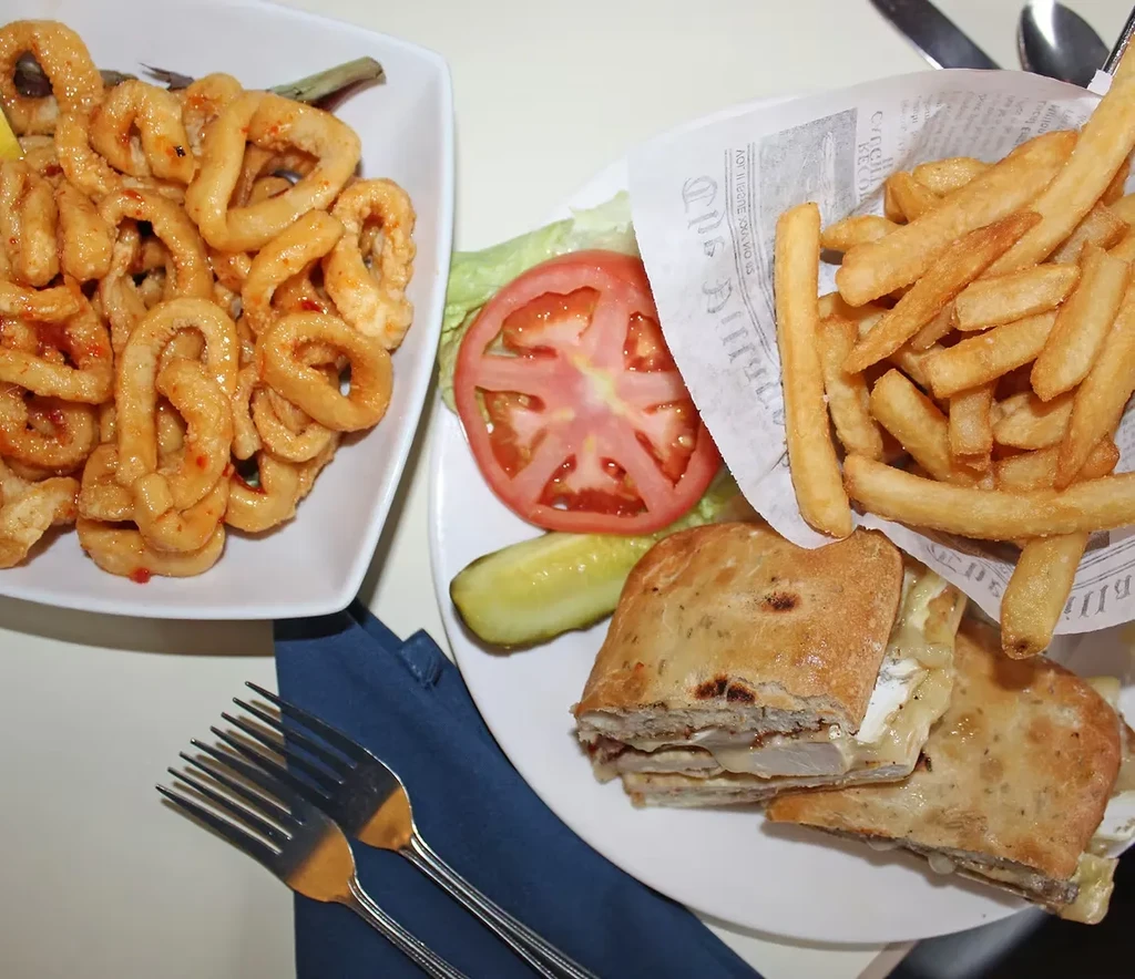 Fried calamari, panini, fries, tomato, and pickle on white plates.