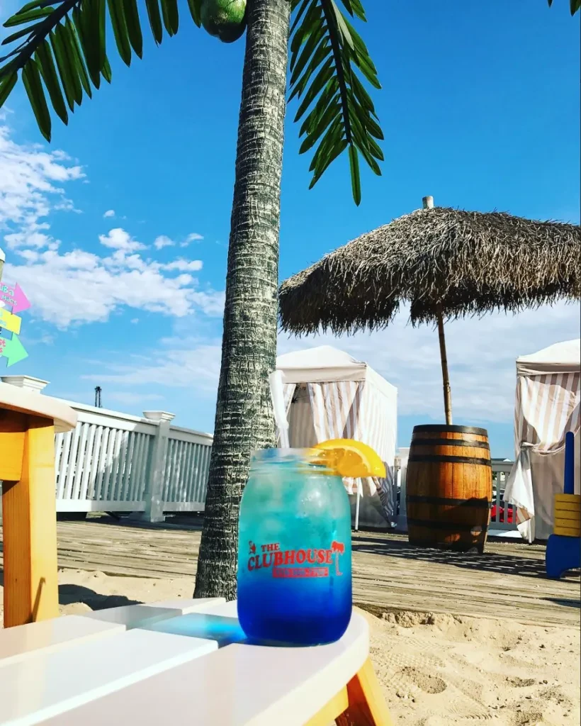 Blue drink on white chair, palm tree, and thatched umbrella on beach.