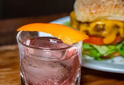 A cocktail with an orange peel and a cheeseburger in the background.