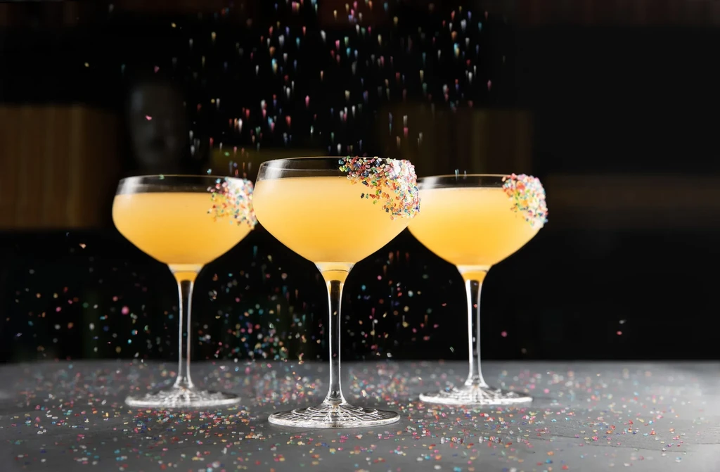 Three yellow cocktails with colorful sprinkles on their rims and falling.