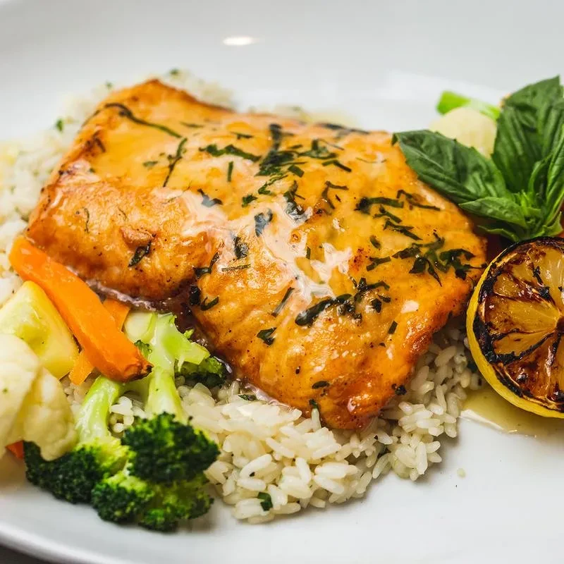 A white plate with a salmon fillet, rice, vegetables, and a lemon.