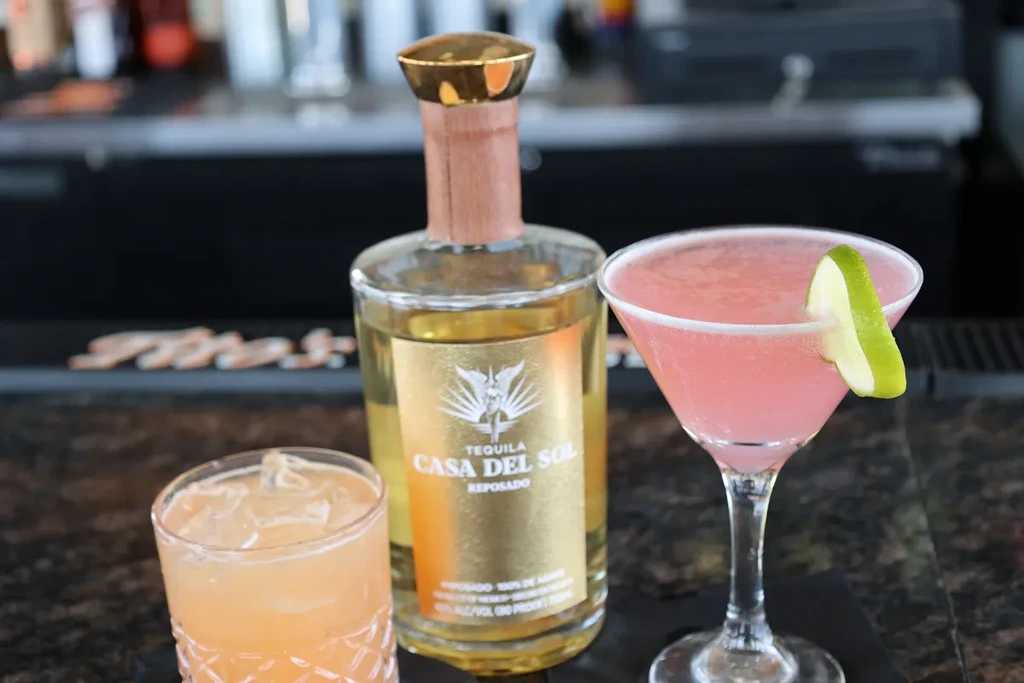 Casa Del Sol tequila bottle, a pink martini, and a light orange cocktail.