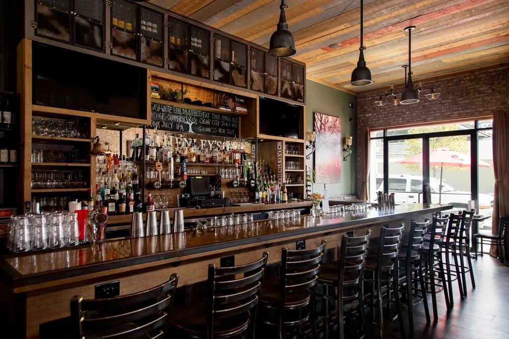 A long wooden bar with stools and shelves of alcohol bottles.