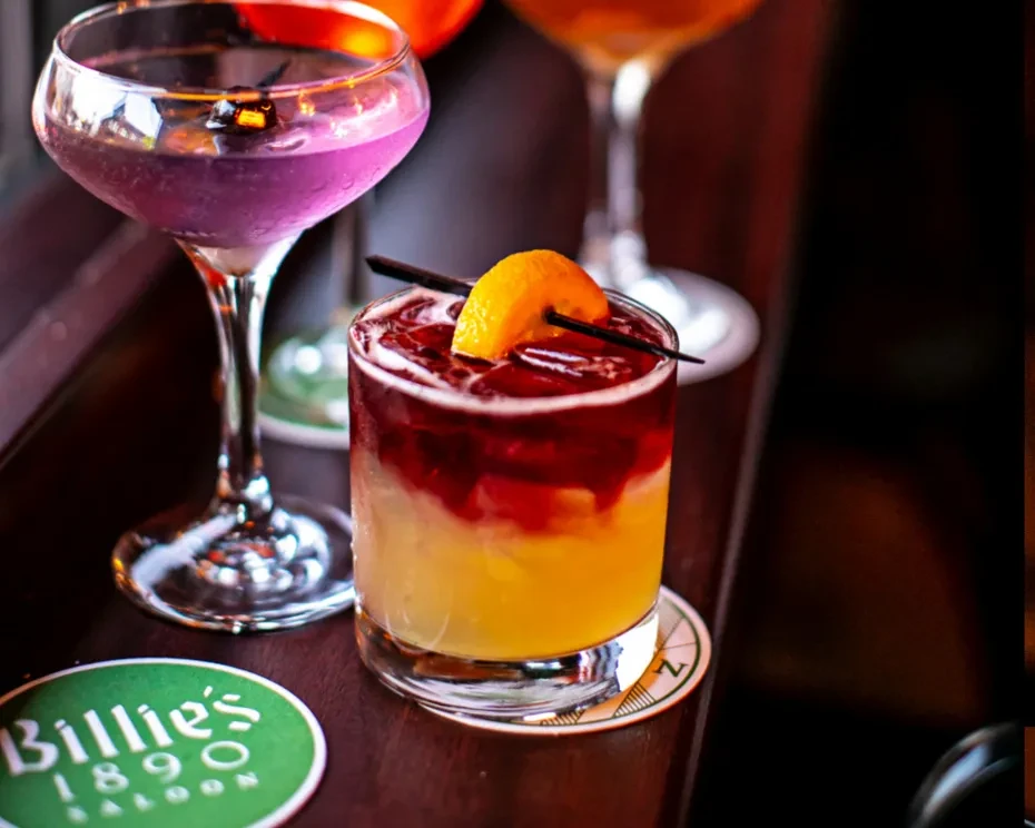 Two cocktails, one purple and one yellow with red, on a bar.