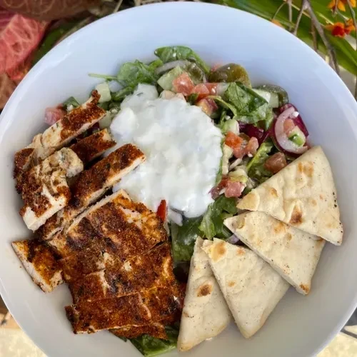 A white bowl of grilled chicken, salad, yogurt sauce, and pita bread.