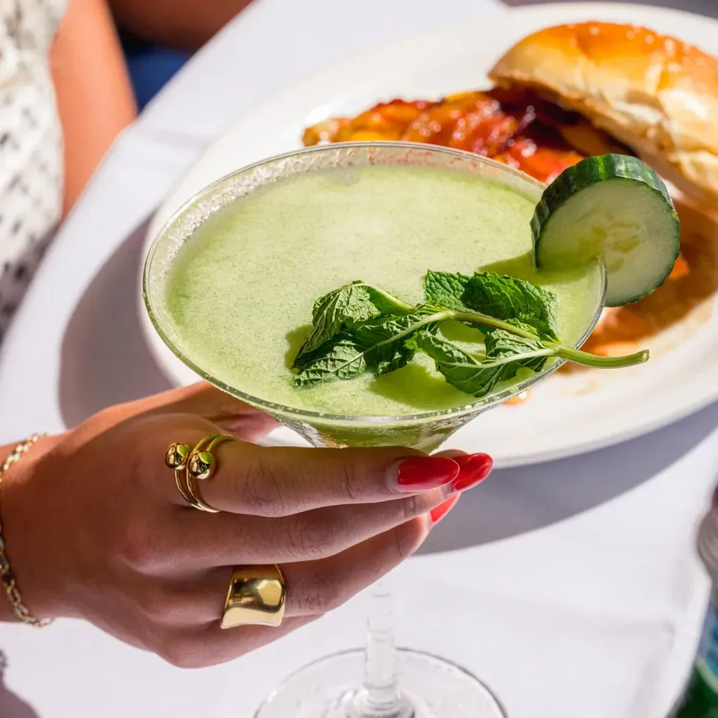 A person holding a green drink garnished with mint and cucumber.