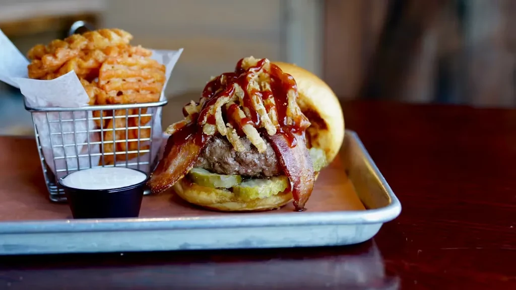 A gourmet burger with waffle fries and sauce on a metal tray.