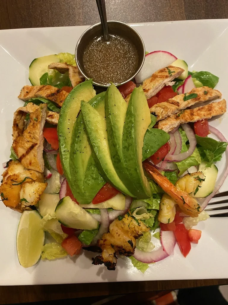 A salad with grilled chicken, avocado slices, shrimp, and lime.