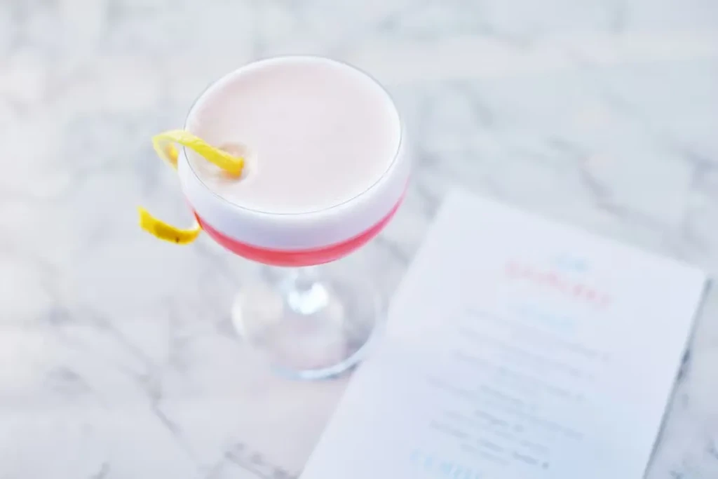 A pink cocktail with foam and lemon twist on a marble surface.
