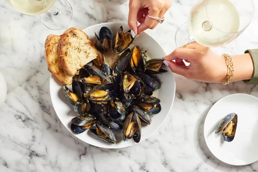 Overhead view of two people eating mussels with bread and white wine.