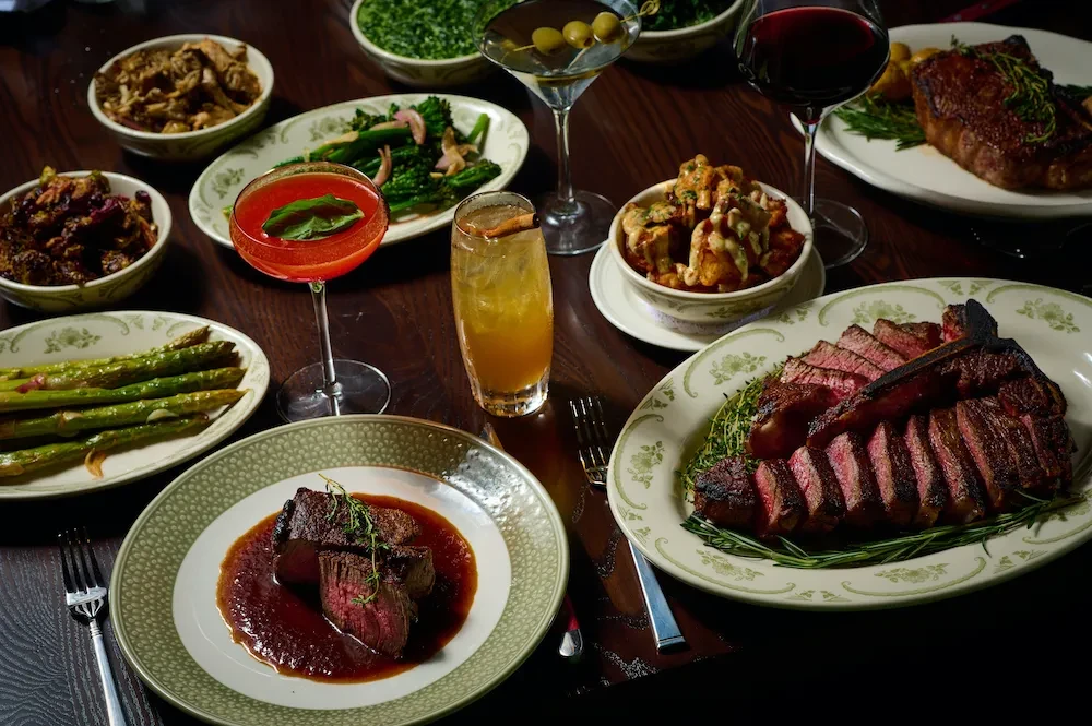 A table laden with multiple plates of steak, side dishes, and cocktails.