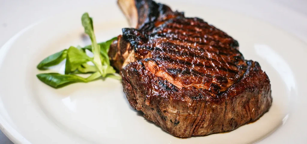A perfectly cooked steak with a bone and greens on a white plate.