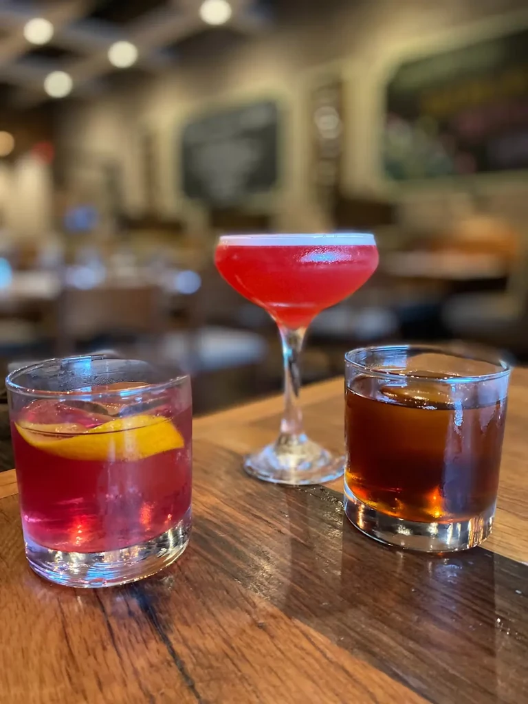 Three cocktails sit on a dark wooden table in a dimly lit bar.