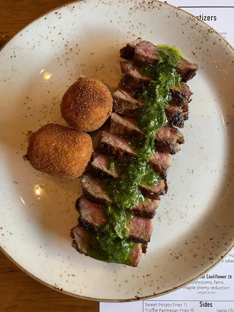 Sliced steak with green sauce and two fried croquettes on a white plate.