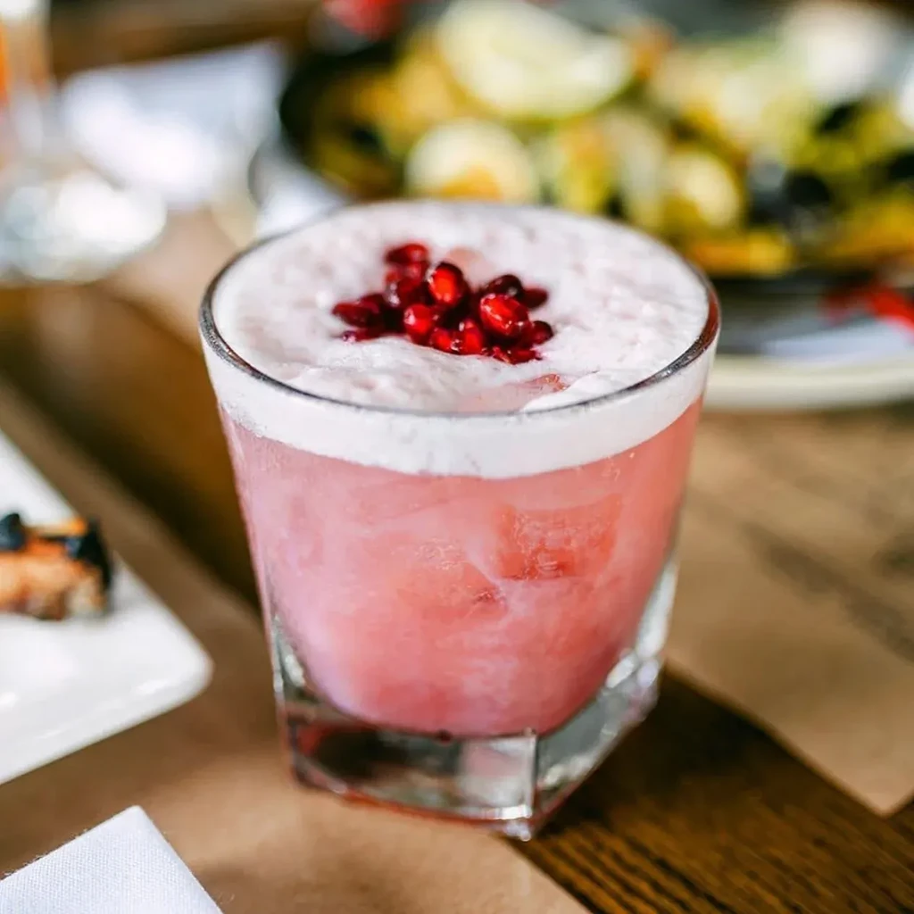 A pink cocktail with a white foam topping and pomegranate seeds.