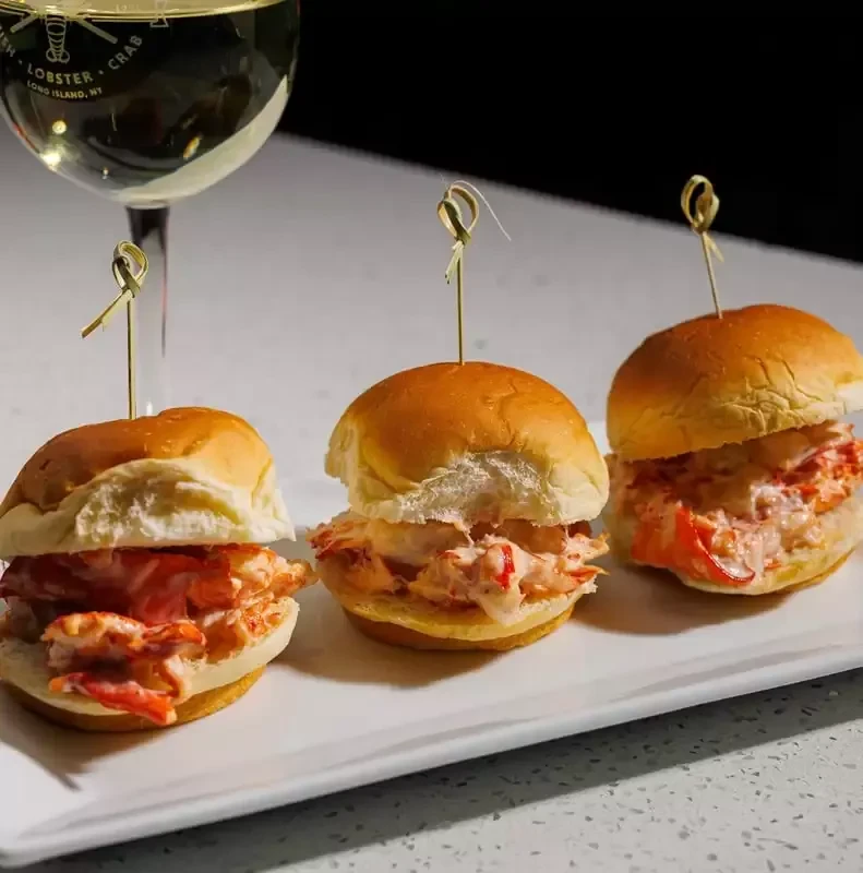 Three lobster sliders on a white plate with a wine glass.