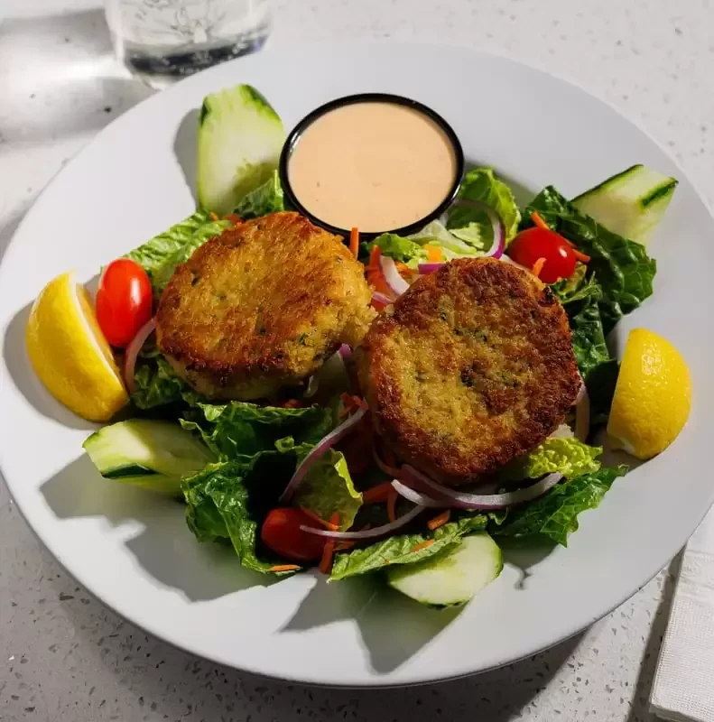 Two crab cakes on a salad with lemons, tomatoes, and sauce.