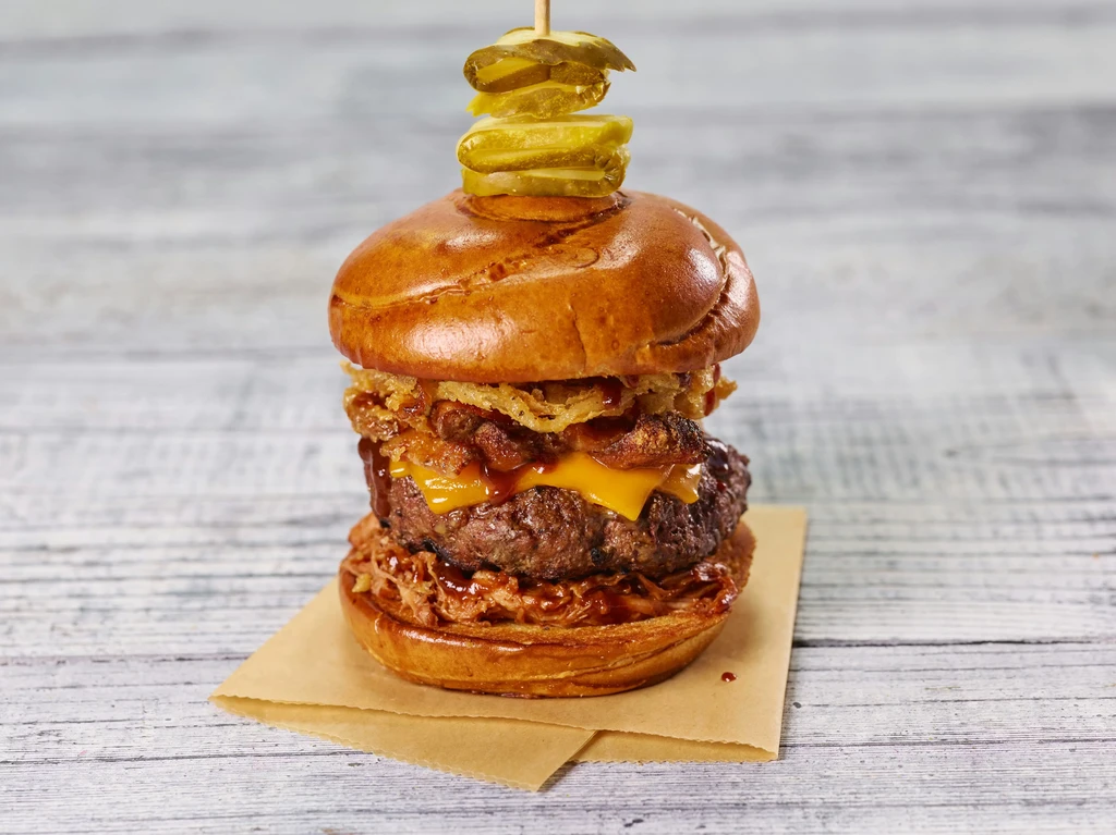 Gourmet burger with beef, cheese, pulled pork, onion rings, and pickles.