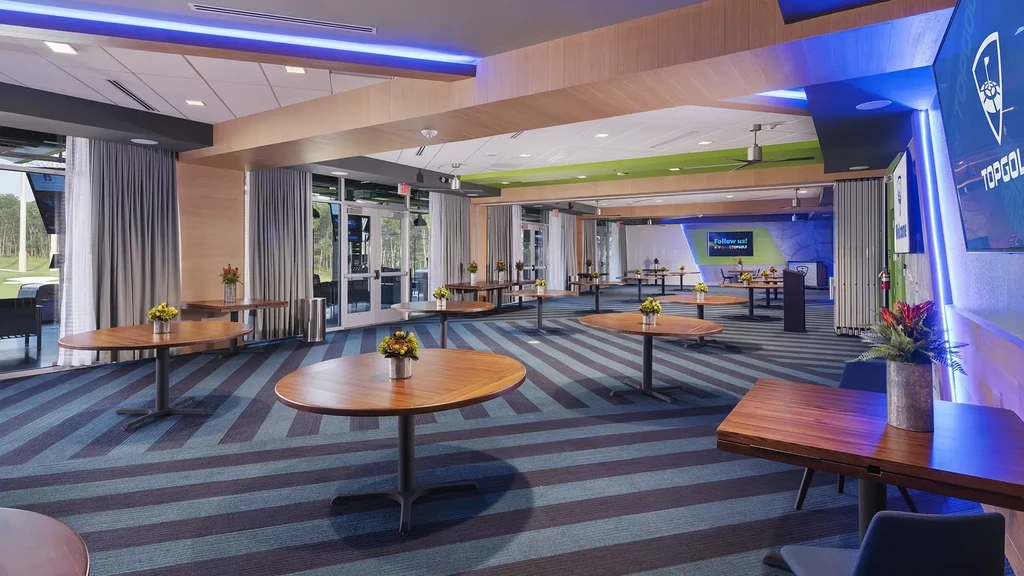 Modern Topgolf event room with tables, striped carpet, and golf range view.