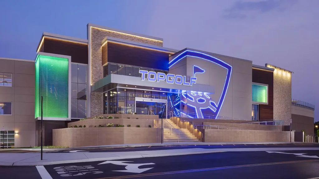 Topgolf building at dusk with blue neon signs and green glowing panel.