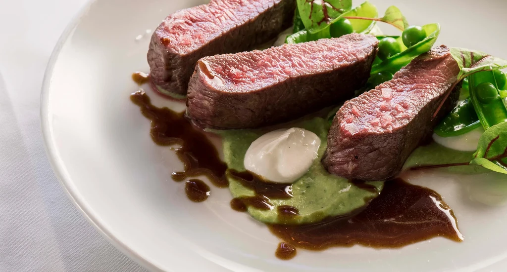 Three medium-rare steak slices with sauce, peas, and green puree on a plate.