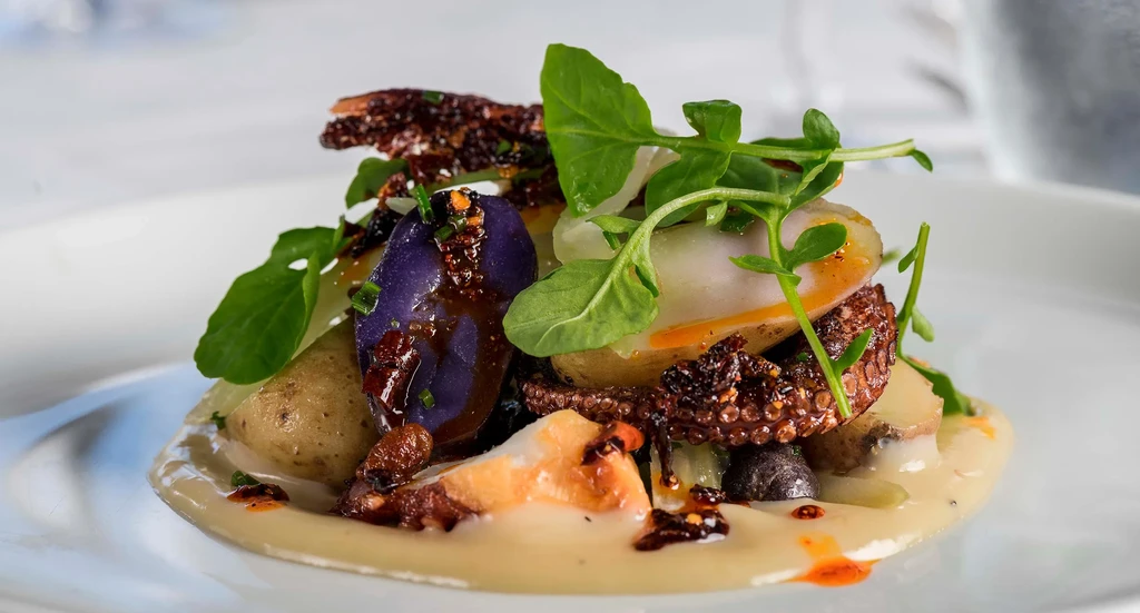 A gourmet plate of octopus, potatoes, purple yam, and greens on a white sauce.