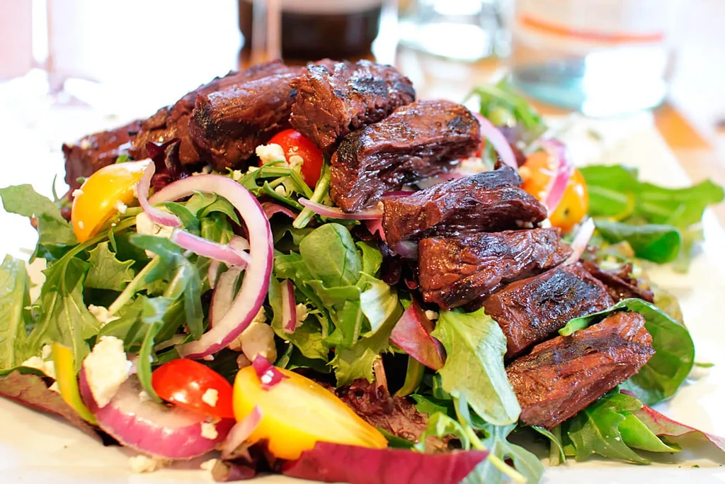Close-up of a grilled beef steak salad with mixed greens, tomatoes, and onions.