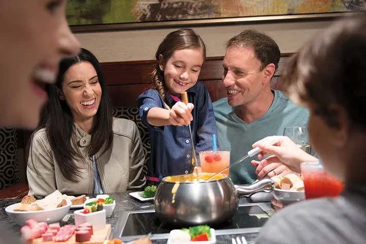 Smiling family at restaurant, a girl stirs a pot of cheese fondue.
