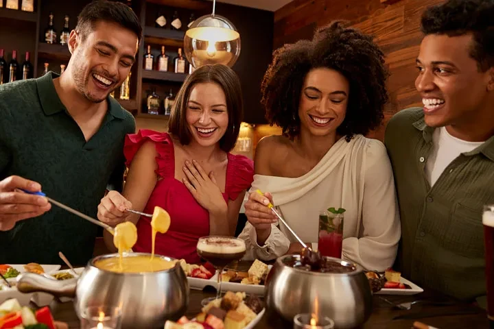 Four smiling people enjoy cheese and chocolate fondue at a modern restaurant.