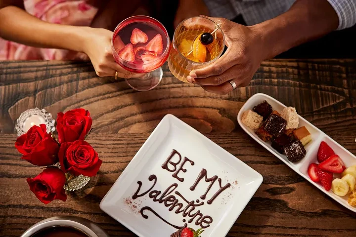 Romantic table with drinks, 'BE MY Valentine' dessert, and treats.
