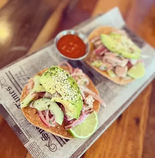 Two tostadas with avocado, onion, and lime, served on newspaper.