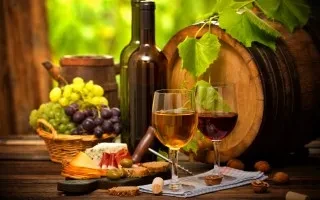 Two wine glasses, bottles, grapes, cheese, and a barrel on a table.