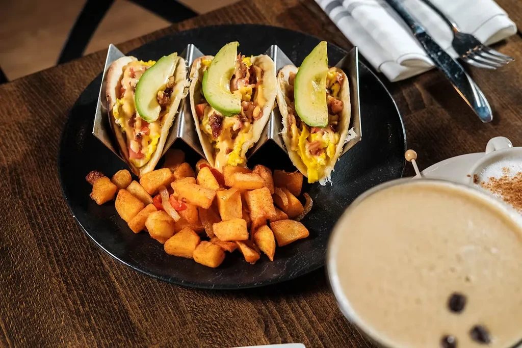 Three breakfast tacos with avocado, scrambled eggs, and bacon on a black plate.