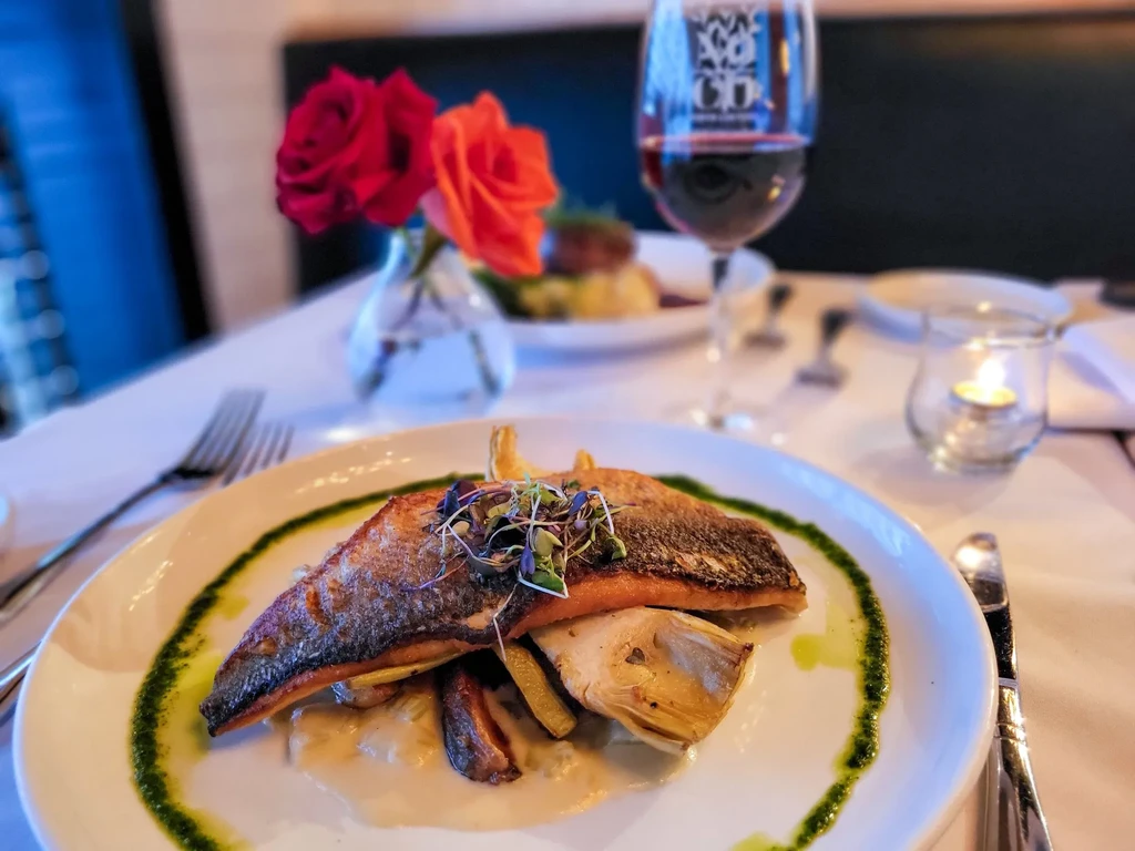 A plated fish entree with herbs, wine, and a candle on a table.
