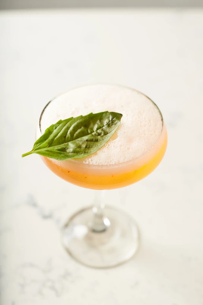A coupe glass with a yellow cocktail, foam, and basil leaf garnish.