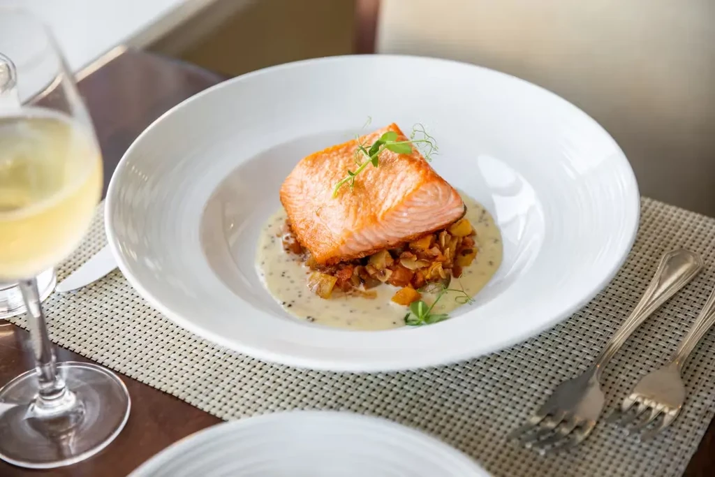 A seared salmon fillet on vegetables with sauce in a white bowl.